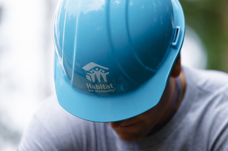 A person wearing a blue Habitat for Humanity hard hat with their head bowed, face obstructed by the hat which features a white HFH logo