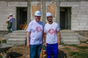 Two volunteers in Habitat T-shirts and hard hats in front of a partly built stone house on a build